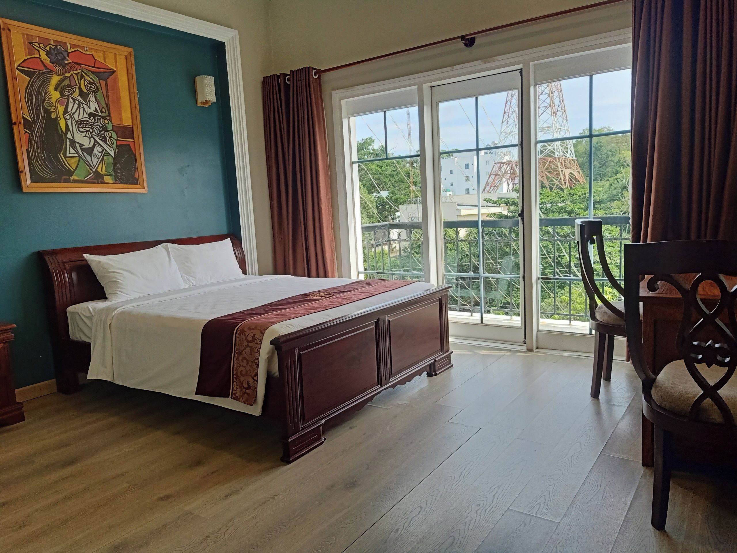 A hotel room with a large bed, a wooden headboard, a painting on the wall, and a balcony with a view of the city.