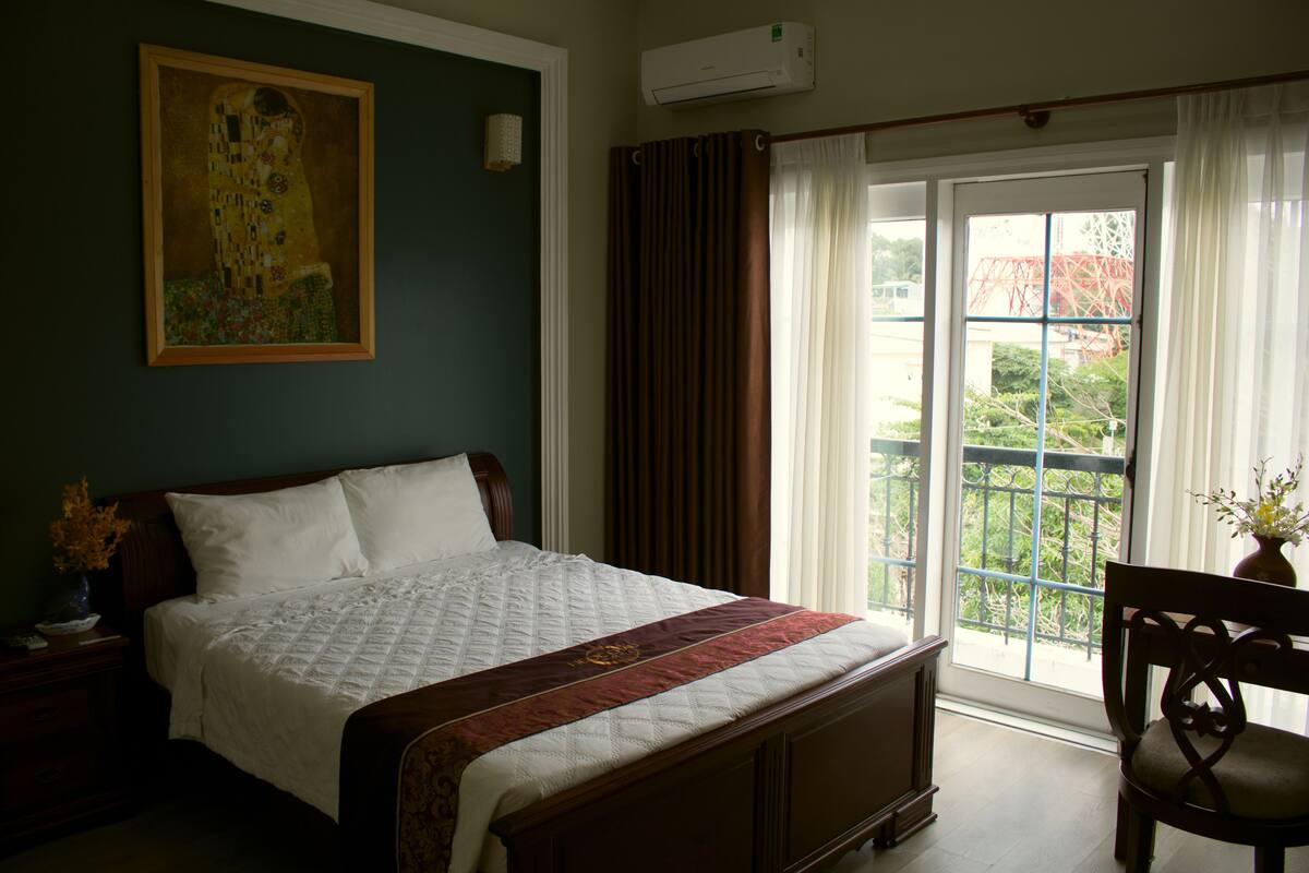 Un dormitorio con una cama grande, una mesita de noche, una silla, un cuadro en la pared y una ventana con cortinas.