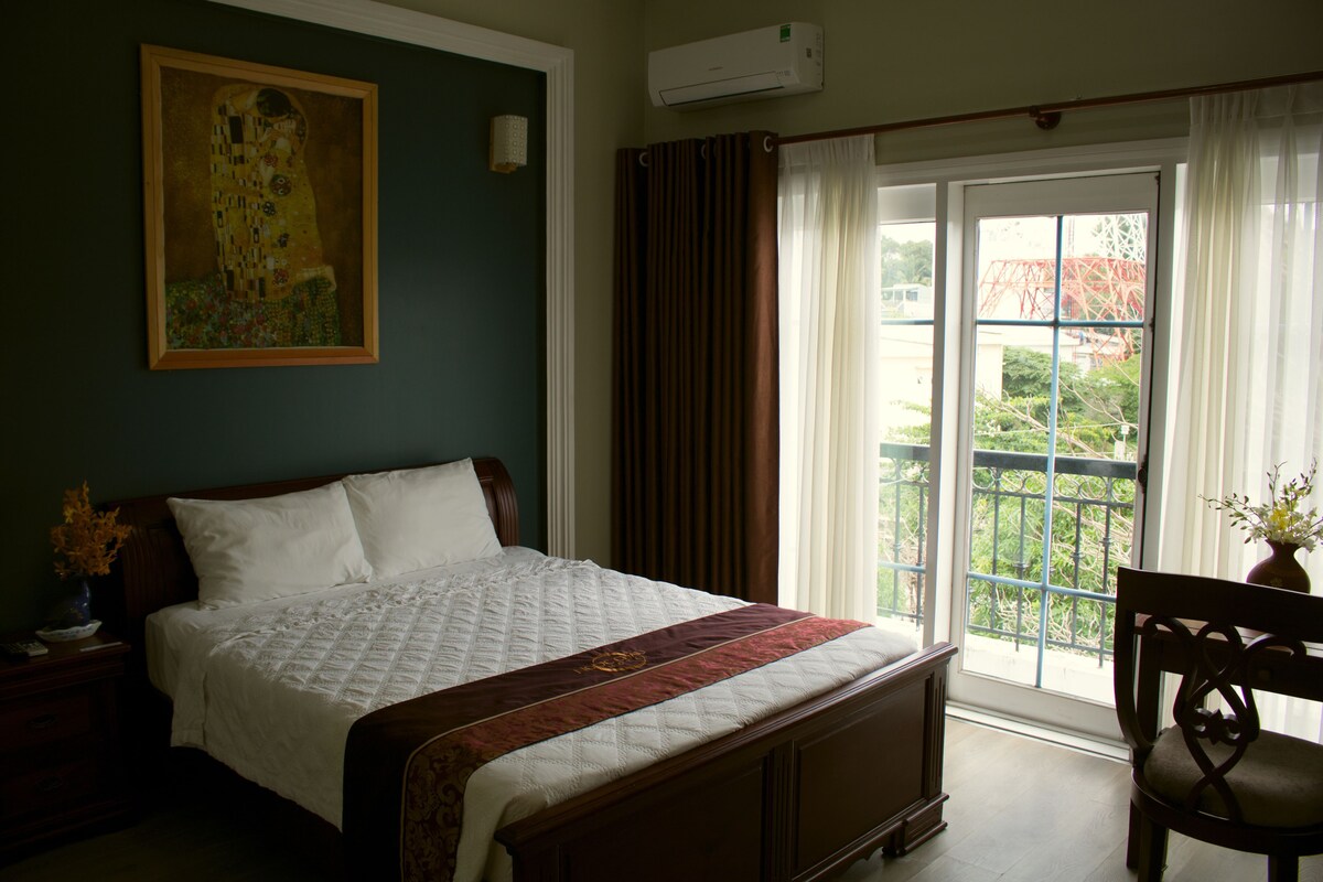 A bedroom with a large bed, a painting on the wall, a window with curtains, and a chair.