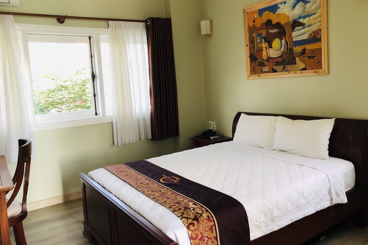 Habitación de hotel con cama, una silla, ventana con cortinas, un cuadro en la pared y un equipo de aire acondicionado.