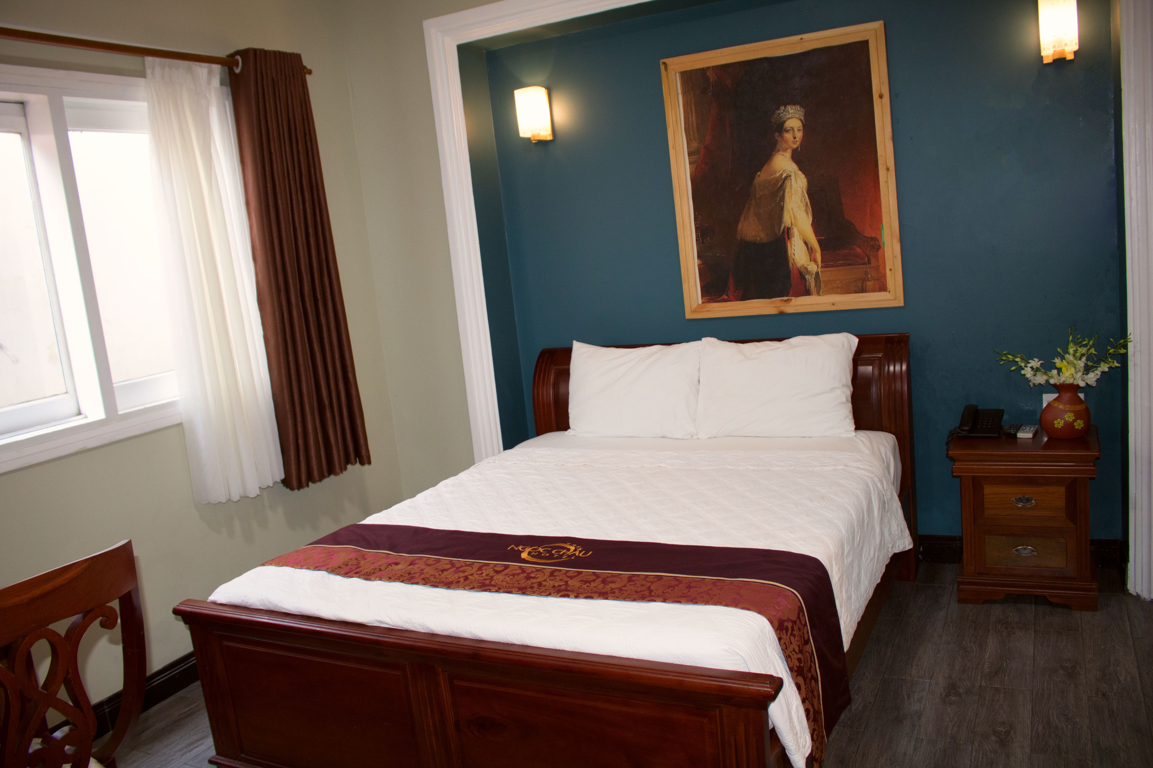 Ein ordentlich bezogenes Bett mit weißer Decke und einem dekorativen Kissen in Burgunder und Gold. Ein Holznachttisch mit einer Vase und einer Lampe steht neben dem Bett. Ein Gemälde hängt an der Wand über dem Bett.
