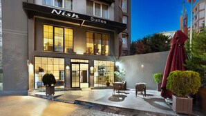 Un hotel anomenat 'Nish Suites' amb façana moderna, grans finestres i zona d'seients a l'exterior.