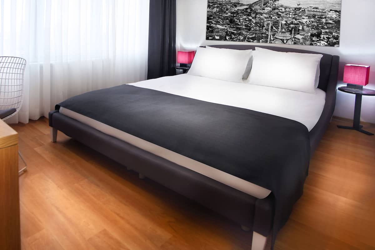 A modern hotel room with a large bed, a desk, a chair, and a cityscape view.