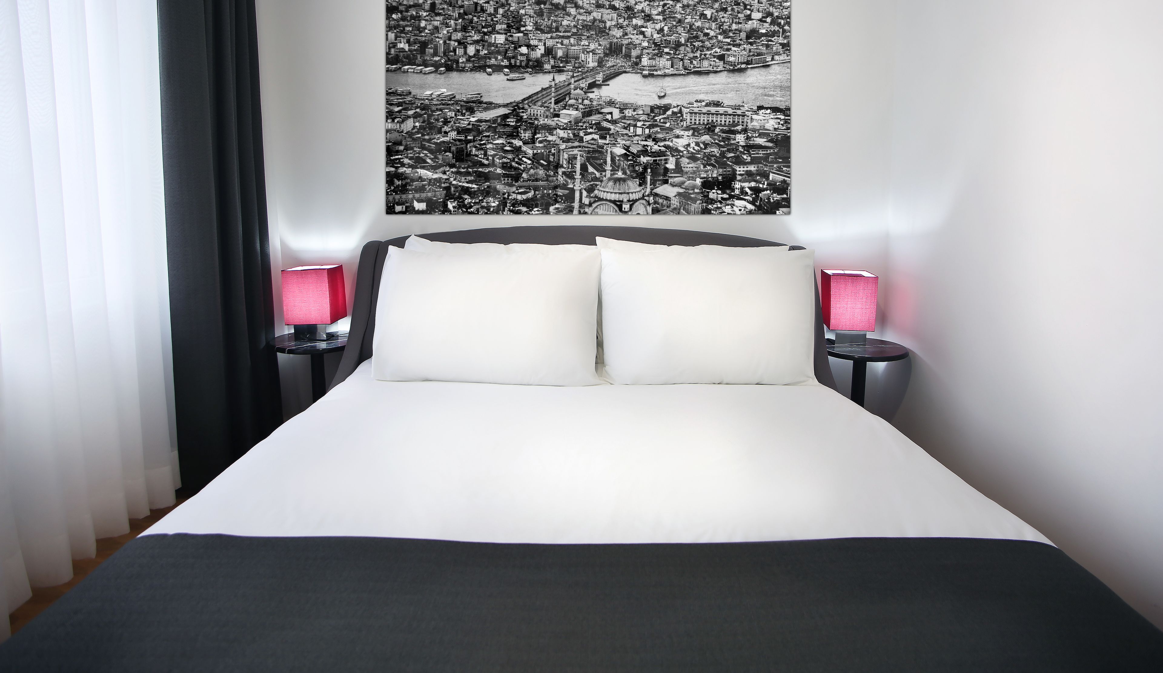 A hotel room with a bed, two bedside tables with lamps, a cityscape artwork on the wall, and a window with curtains.