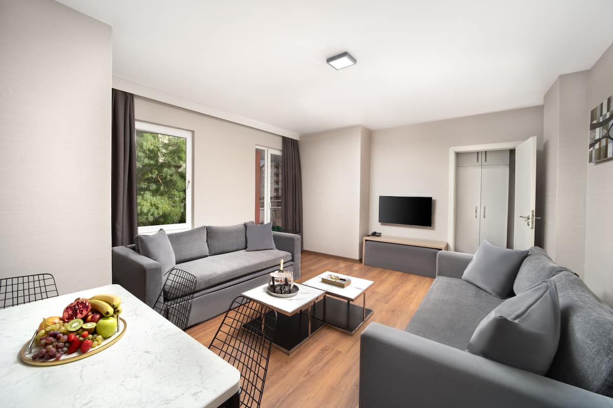 A modern living room with a grey sofa, a dining table with fruit, and a TV on the wall.