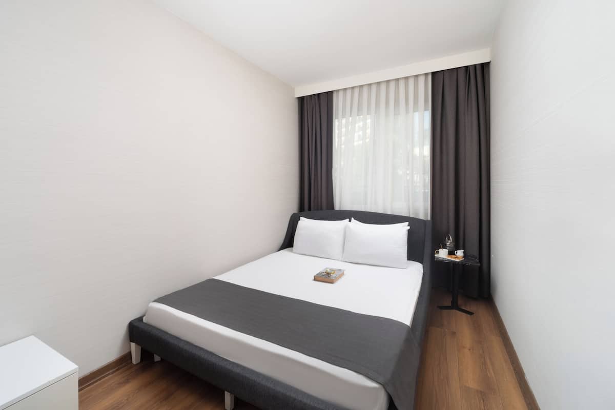 A neatly made bed with a white and gray duvet, a bedside table with a lamp, and a window with dark curtains.