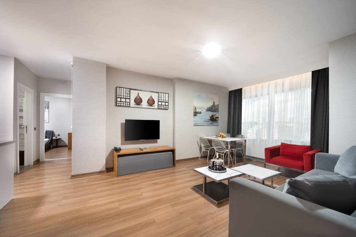 A modern living room with a flat-screen TV, a dining area with a table and chairs, a red sofa, and a grey armchair.