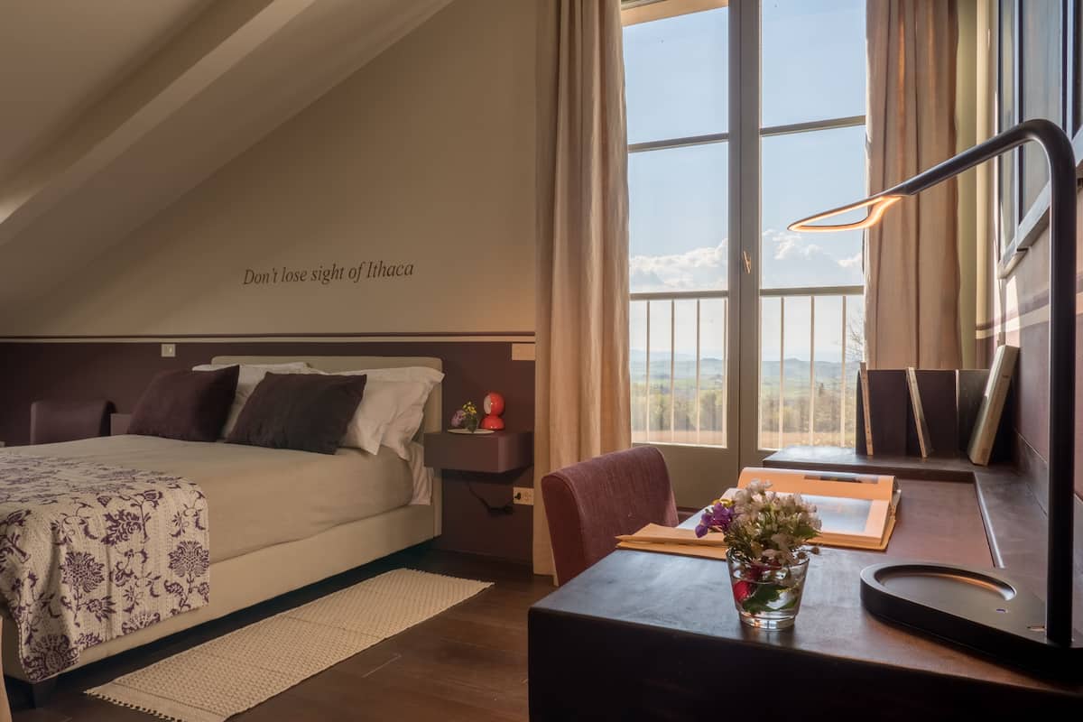 A hotel room with a bed, a desk, a chair, a nightstand, a lamp, and a window with a view.