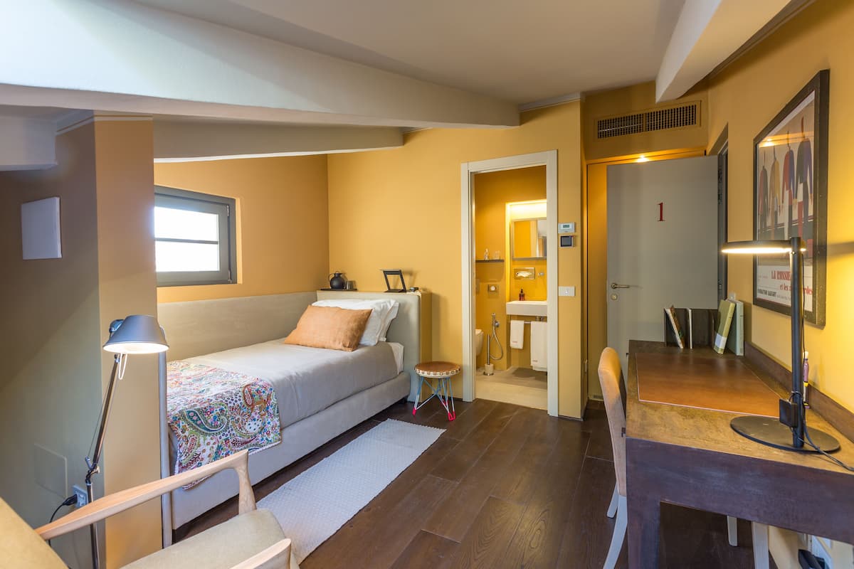 A compact hotel room with a bed, bedside table, lamp, and a small stool.