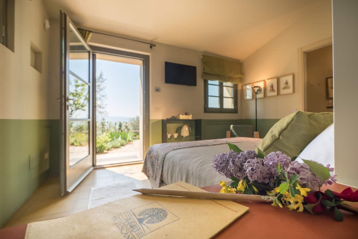A bedroom with a bed, a desk with a laptop, and a window with a view of greenery.