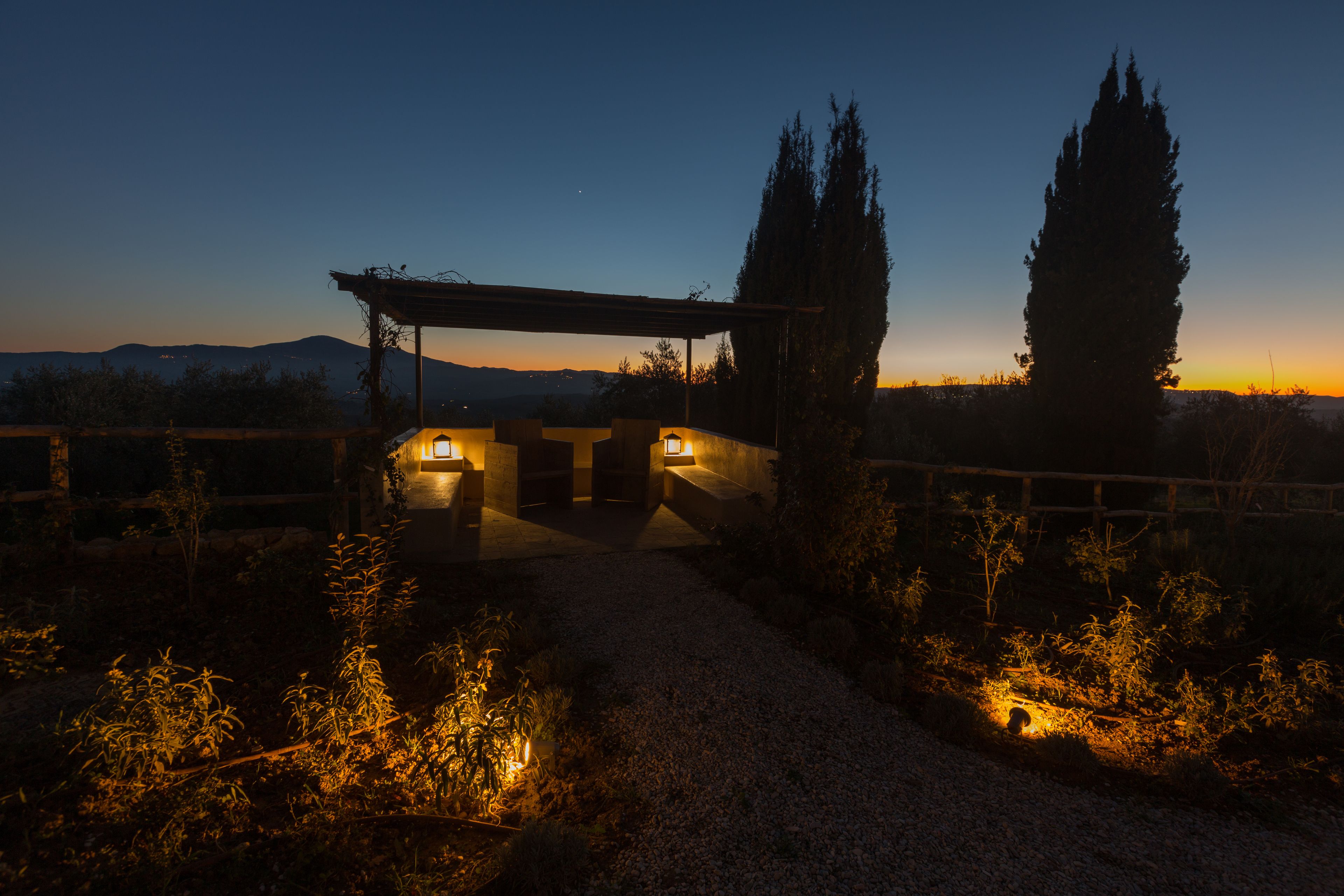 A nighttime view of a rustic outdoor area with a wooden pergola, a gravel path, and tall cypress trees.