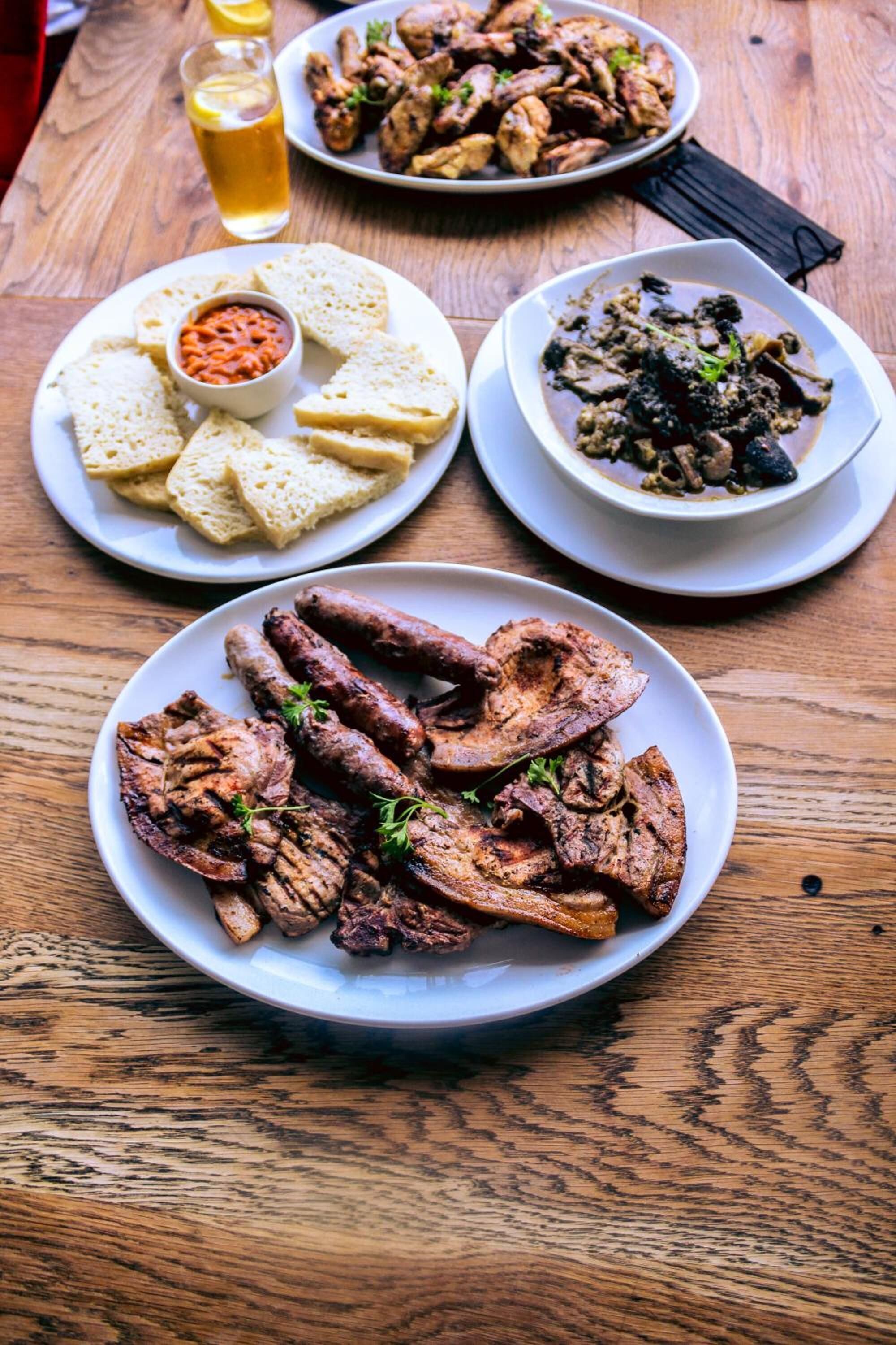 A wooden table with four plates of food, including grilled meat, bread, and a bowl of sauce.