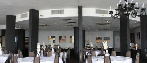 Un vaste espace dédié à la salle à manger, avec des tables et des chaises rondes, un lustre et un plafond haut doté d’un éclairage encastré.