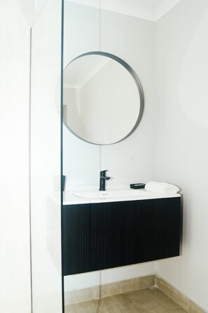 Un baño moderno con ducha de vidrio, un lavabo blanco y una encimera negra, y un espejo redondo.