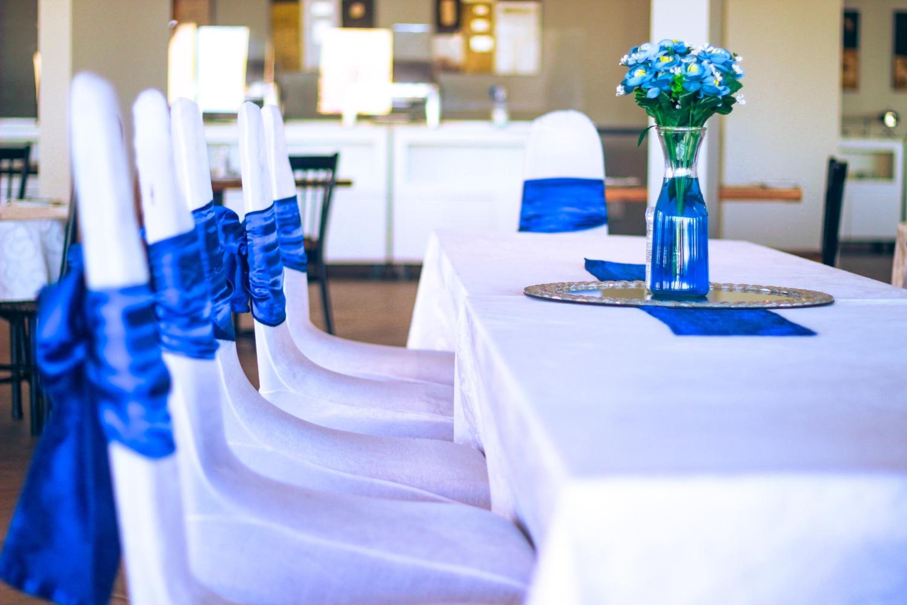 A dining area with white chairs and blue sashes, a table with a blue vase, and a kitchen in the background.