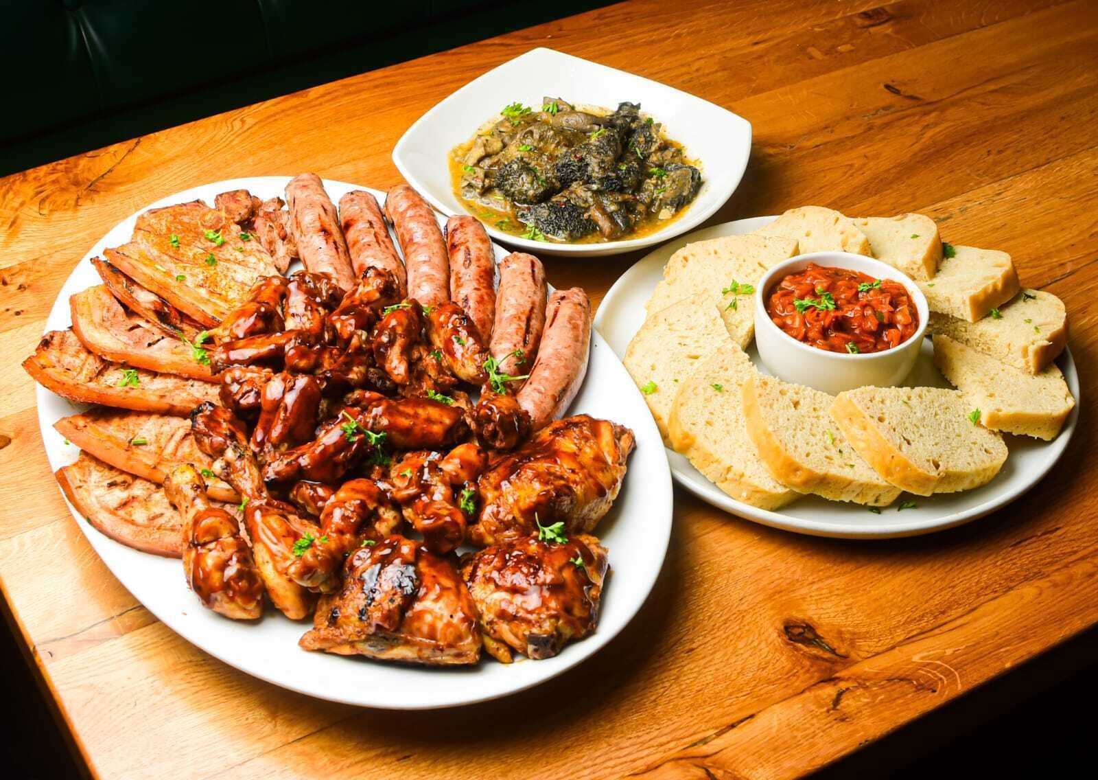 A wooden table with a plate of grilled chicken wings, a bowl of mushroom dish, and slices of bread with a dipping sauce.