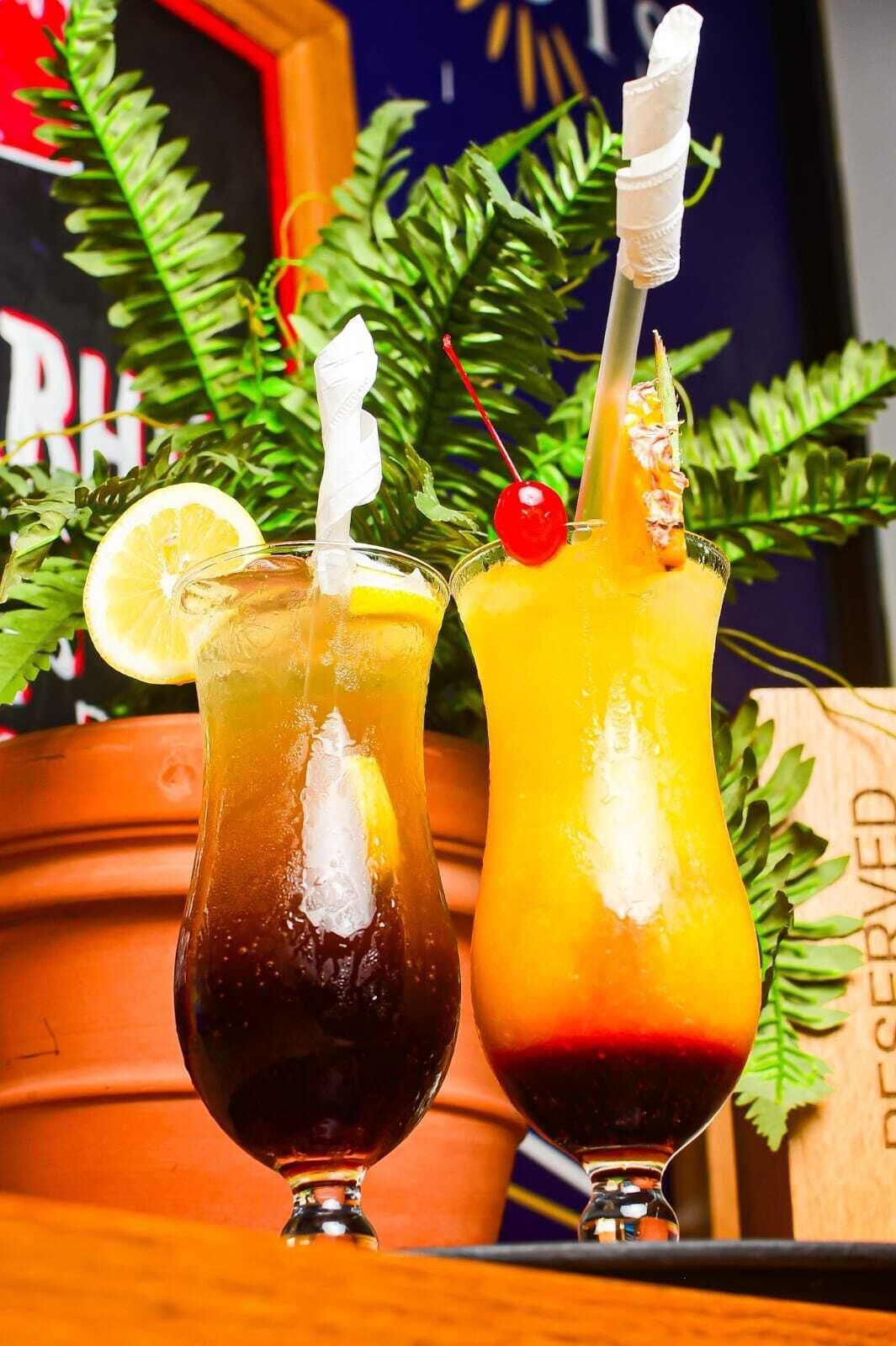 Two cocktails in glasses with ice, garnished with a lemon slice and a cherry, set against a backdrop of green foliage and a wooden barrel.