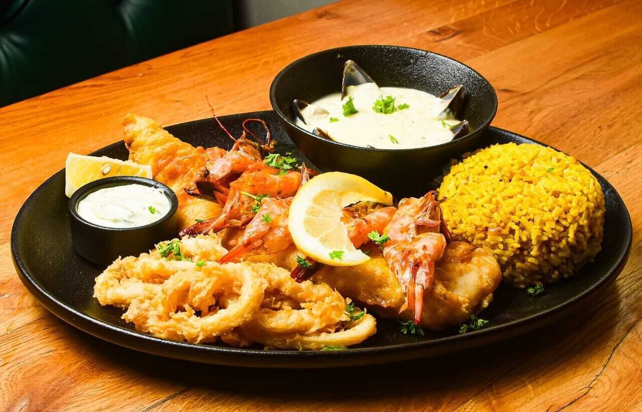 Ein Fisch- und Meeresfrüchtegericht mit Garnelen, Calamari und Reis, serviert auf einem schwarzen Teller mit Zitronenspalten und einer Sauce.