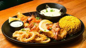 Ein Fisch- und Meeresfrüchtegericht mit Garnelen, Calamari und Reis, serviert auf einem schwarzen Teller mit Zitronenspalten und einer Sauce.