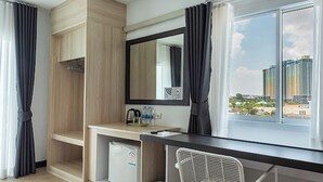 A modern hotel room with a wooden wardrobe, a small refrigerator, a desk with a mirror, and a view of buildings through a window. - Le Nara Residence (Pattaya)