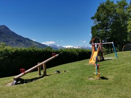 Un parque infantil con un sube y baja y un tobogán, con montañas y cielos despejados de fondo.