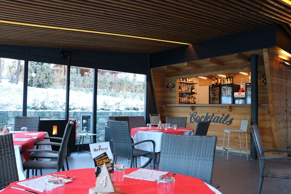 An outdoor dining area with wicker chairs and red tablecloths, a bar area with a 'Cocktails' sign, and a view of trees outside. - Lazy Bee Camping Village (Quart)