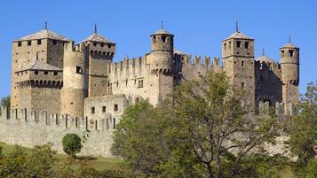 Un château de pierre historique, doté de nombreuses tours et de créneaux, entouré d’un mur de pierre et de verdure.