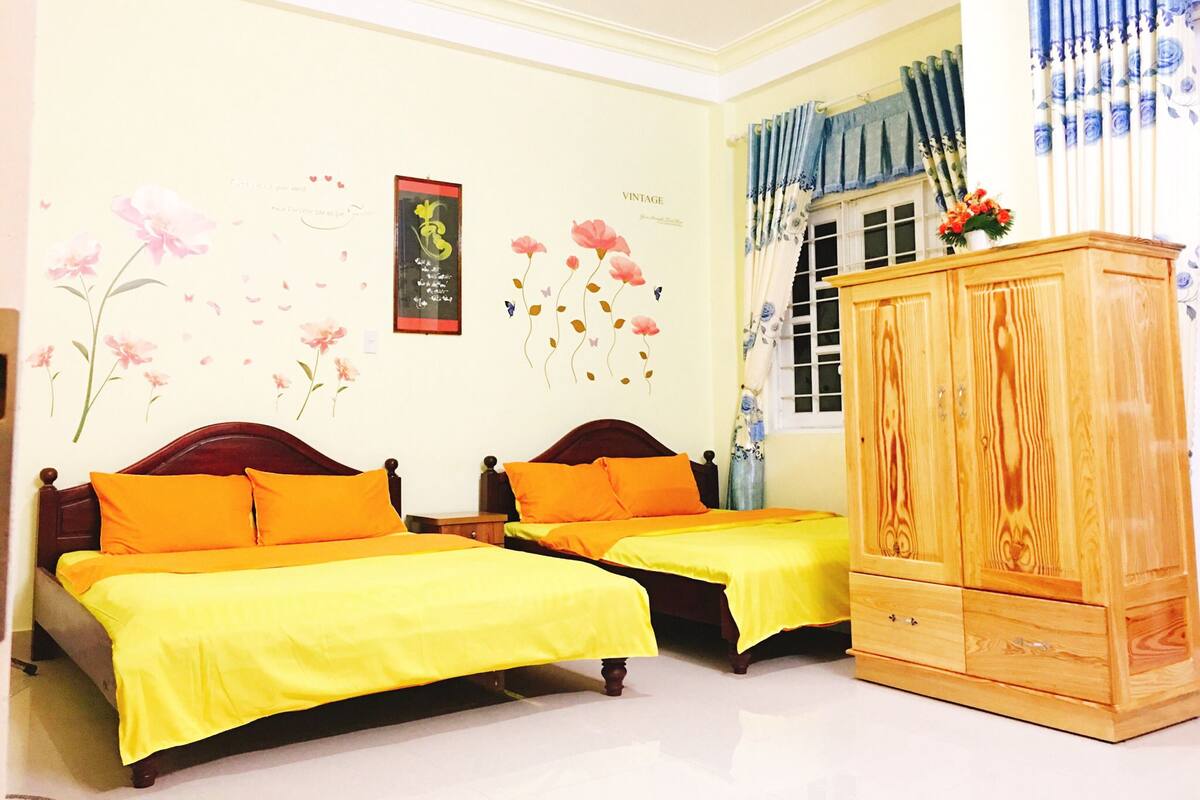 A room with two beds, each with an orange bedspread and pillows, a wooden cabinet, and floral wall decorations.