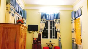 A room with a wooden cabinet, a television, a small table with two red stools, a window with blue curtains, and a door with a yellow gate. - Pensee Guesthouse (Lam Vien - Da Lat)