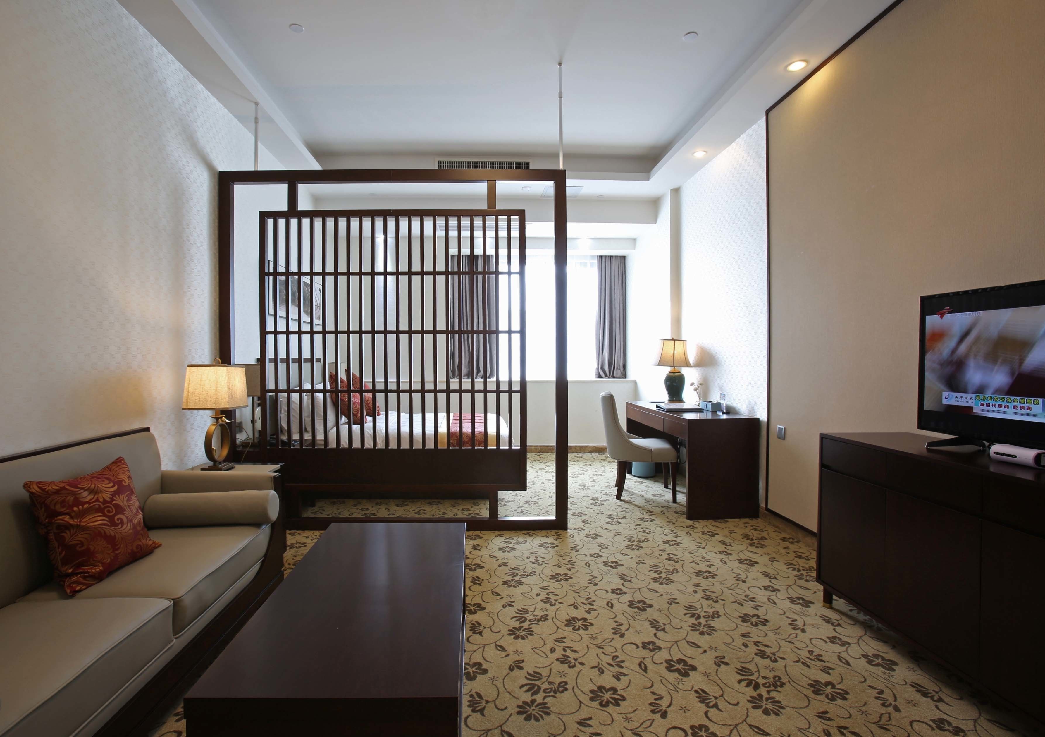 A hotel room with a sofa, a coffee table, a TV, a desk with a lamp, and a decorative screen.
