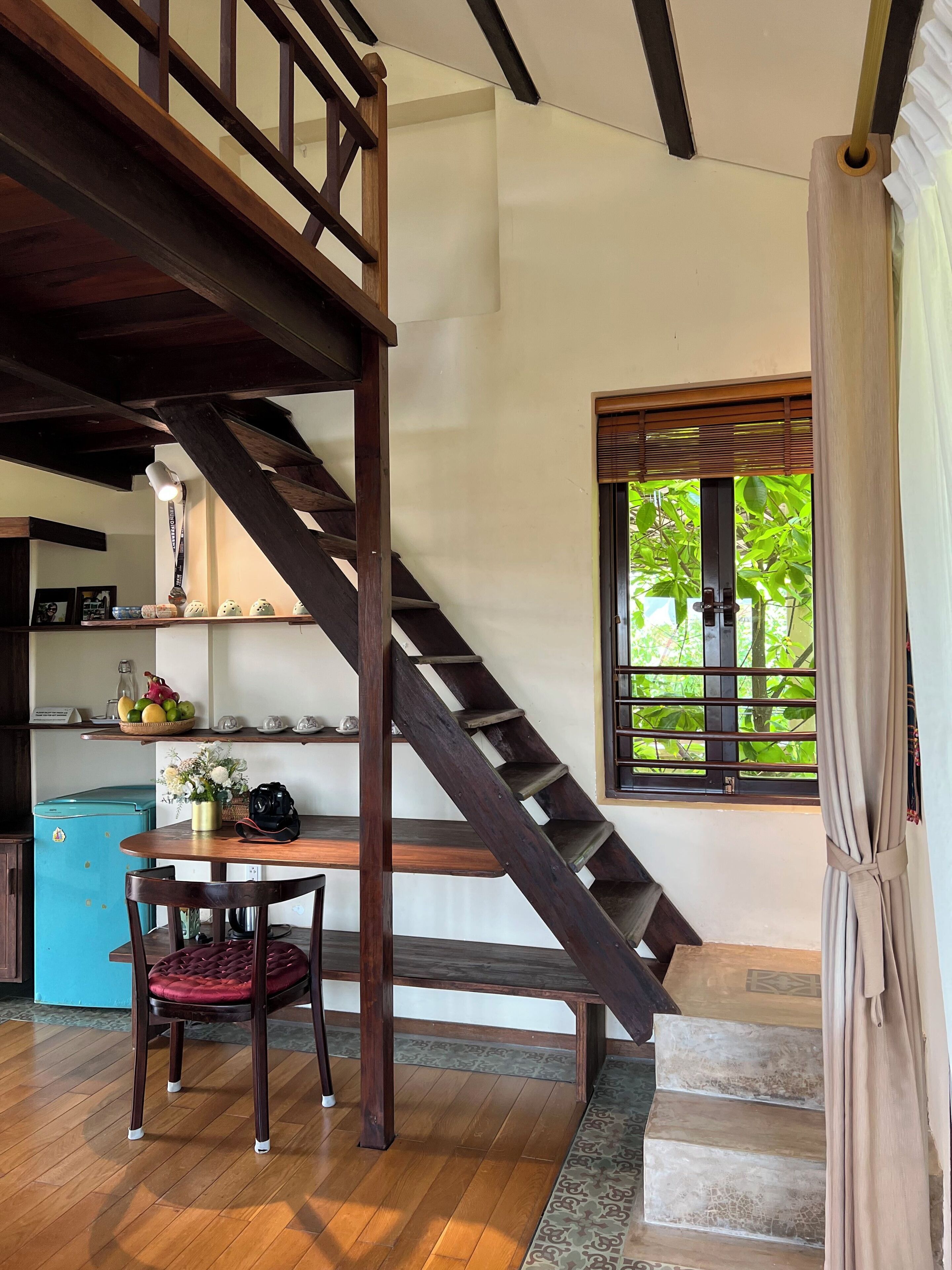 A wooden staircase leading to an upper level, a dining area with a table and chairs, a blue refrigerator, and a window with a view of greenery.