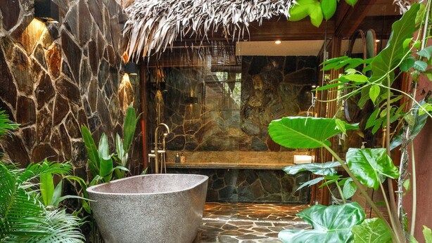 A stone-walled bathroom with a large stone bathtub, surrounded by lush tropical plants. - An Villa Hoi An (Da Nang)