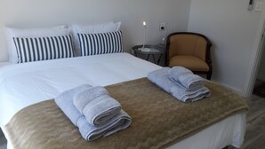Une chambre d’hôtel avec un lit, deux oreillers, une chaise, une petite table avec une lampe et deux serviettes pliées posées sur une couverture beige.