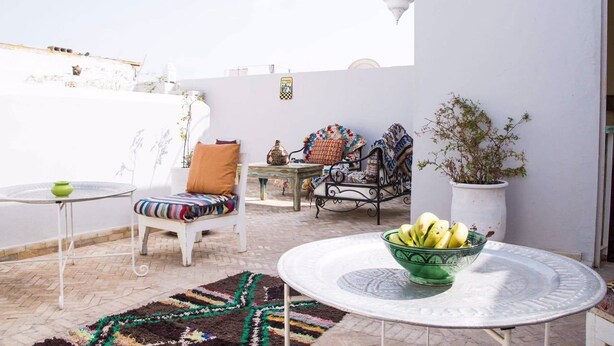 A terrace with a white chair, a small table with a bowl of fruit, a patterned rug, and a potted plant. - Riad el Marah (Marrakech)
