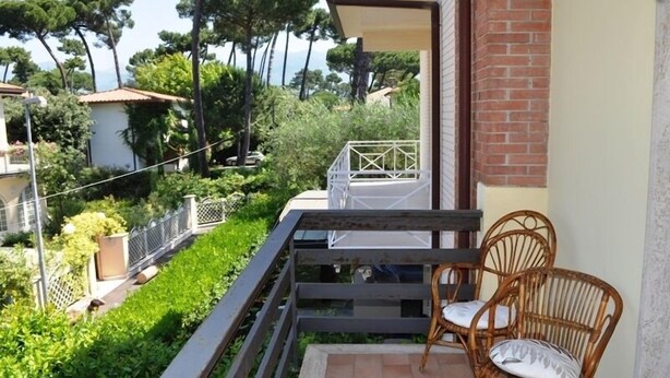 A balcony with a wooden railing, wicker chairs, and a view of a lush garden and distant buildings. - Villa Fiumetto (Pietrasanta)