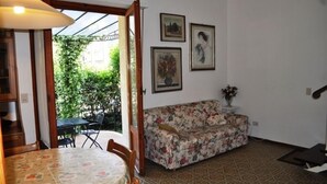 A room with a floral sofa, a dining table with chairs, and a small side table with a vase. There are framed pictures on the wall and a door leading to an outdoor area with a table and chairs. - Villa Fiumetto (Pietrasanta)