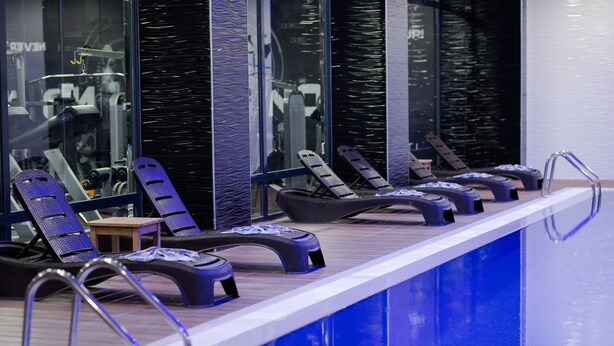 Zona de piscina con sillones de descanso y una pequeña mesa, rodeada de paredes de vidrio que reflejan equipo de gimnasio.