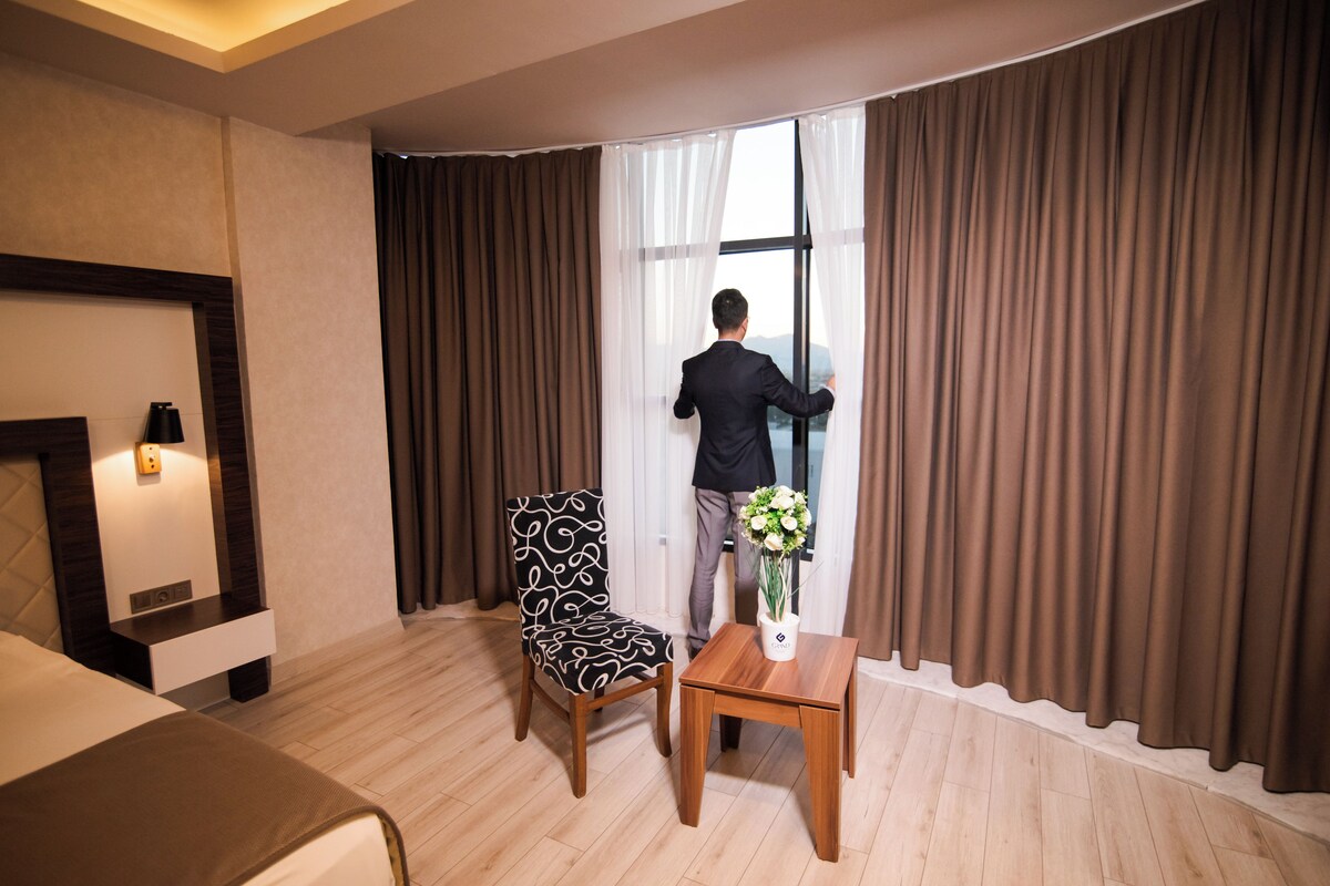 A hotel room with a bed, a chair, a small table with a vase of flowers, and a window with curtains.