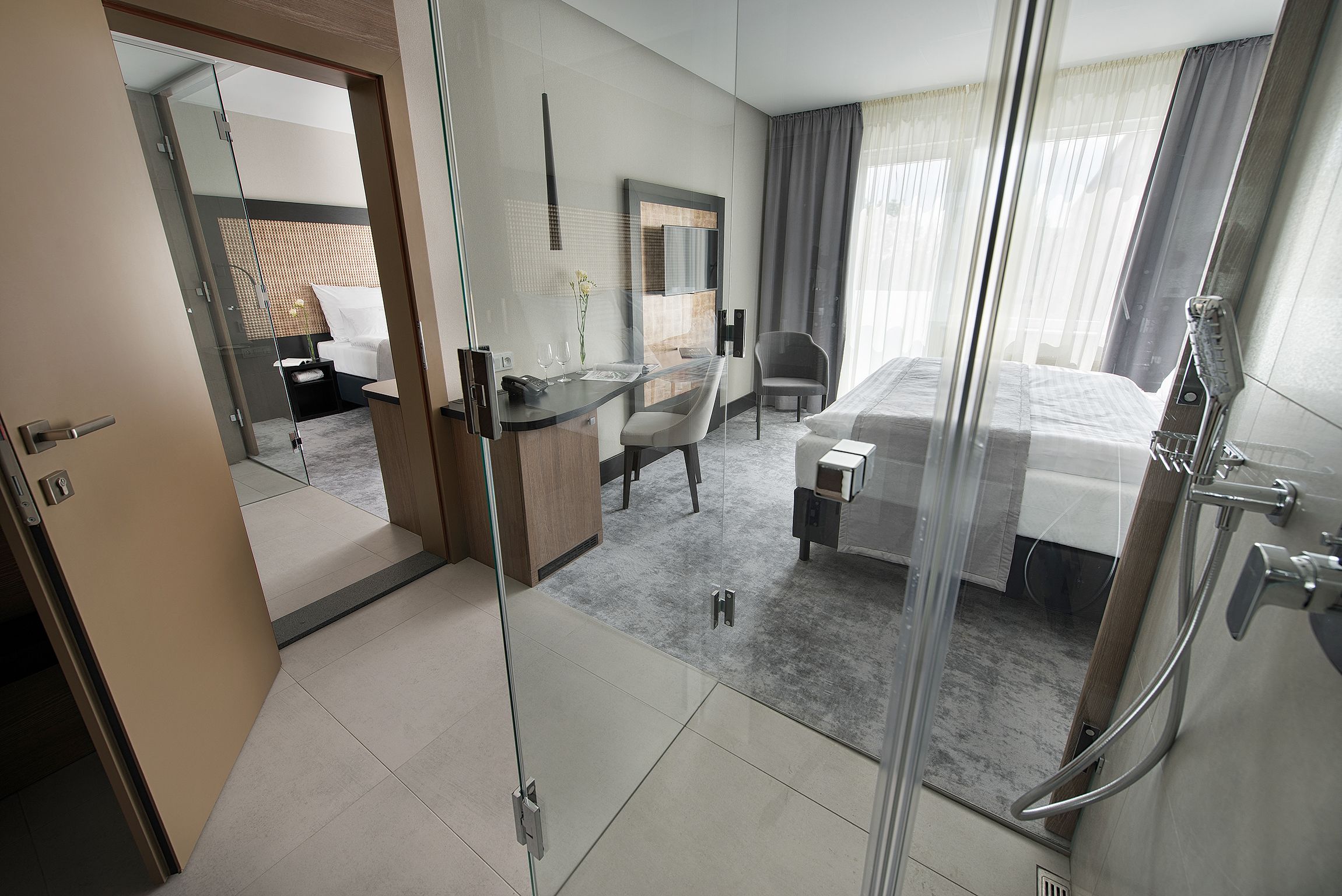 Habitación de hotel moderna con separador de vidrio, cama, escritorio con silla, zona de ducha y vistas a la ciudad.