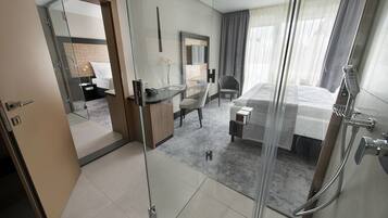 Habitación de hotel moderna con separador de vidrio, cama, escritorio con silla, zona de ducha y vistas a la ciudad.