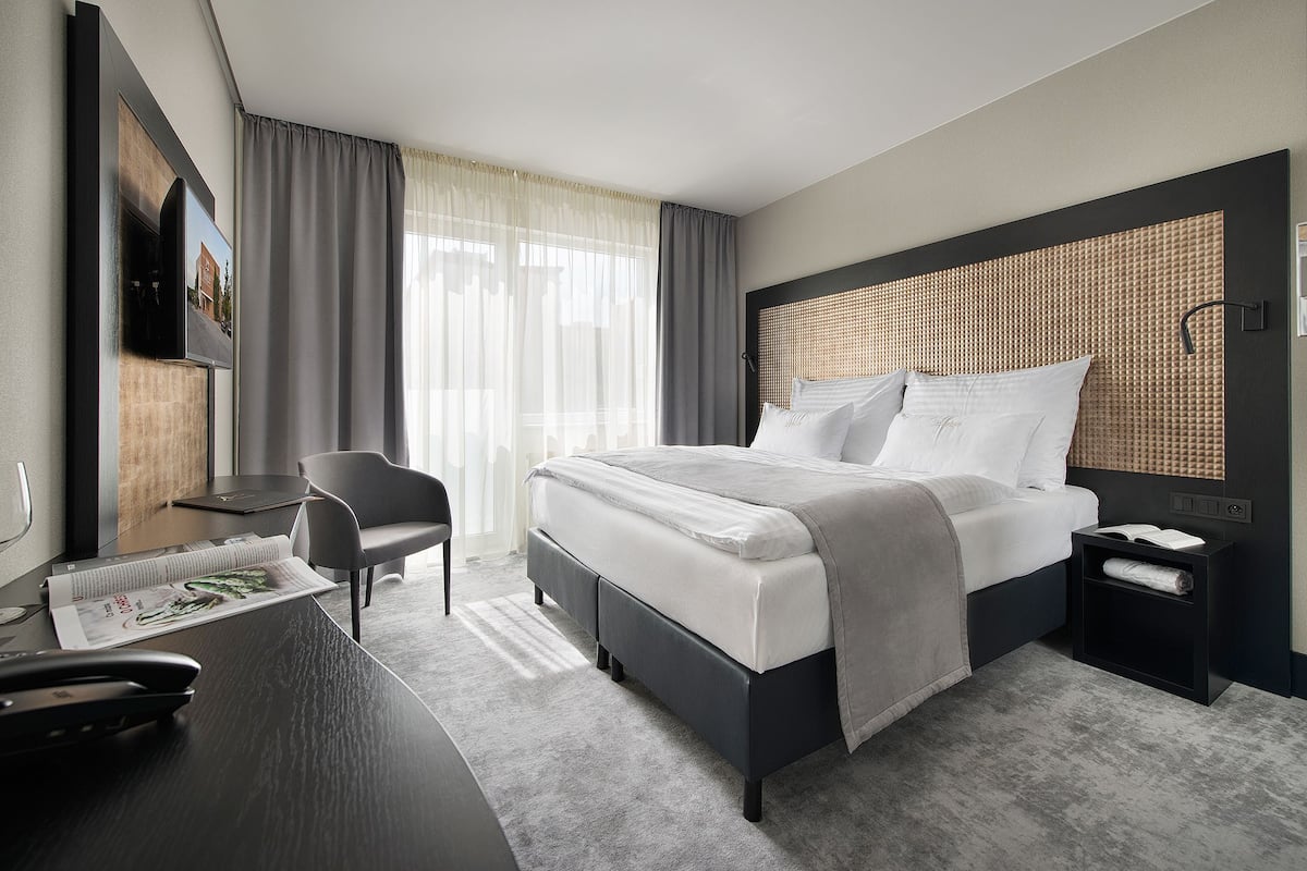 A modern hotel room with a large bed, a desk with a chair, a flat-screen TV mounted on the wall, and a large window with sheer curtains.