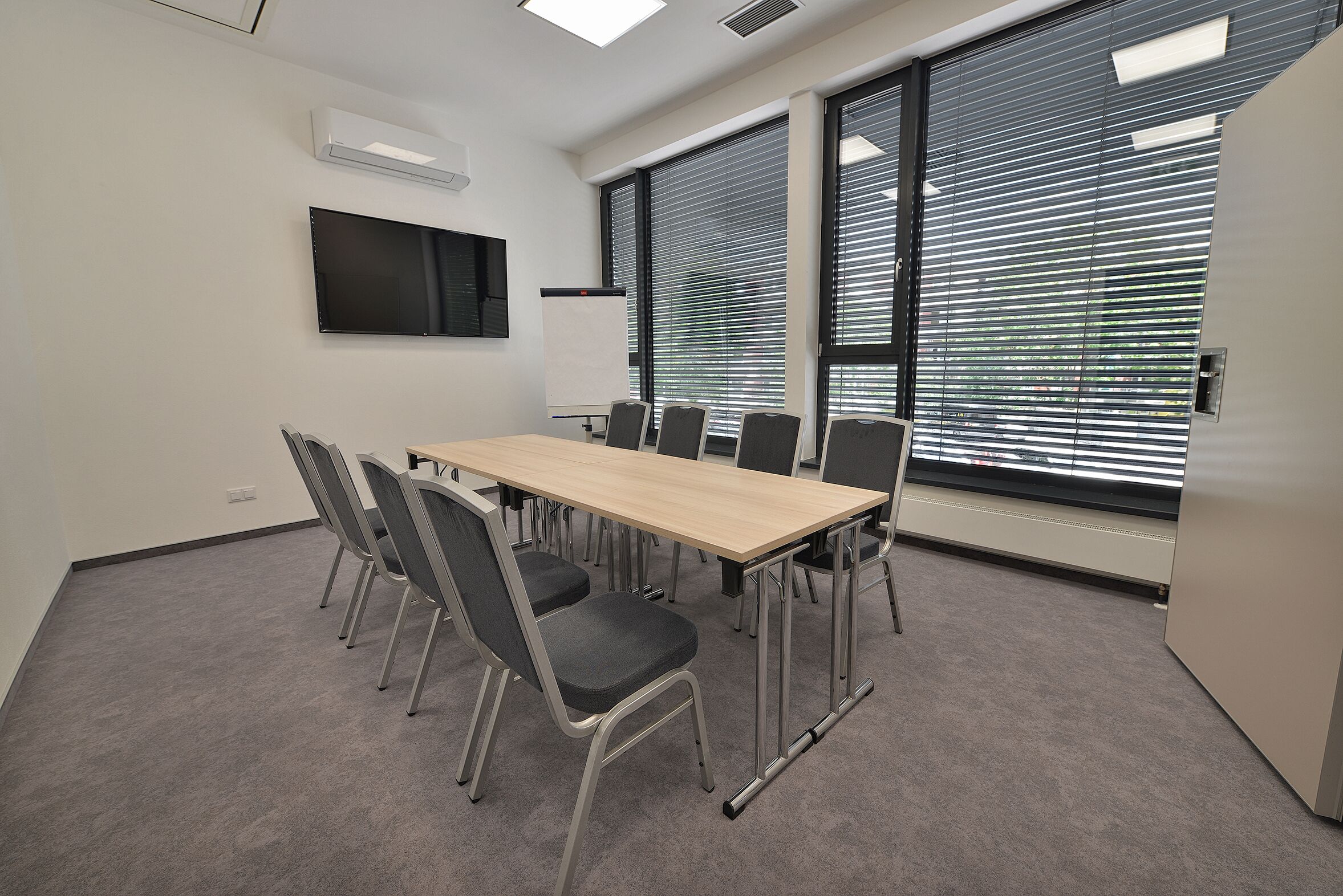 A conference room with a long table, six chairs, a flat-screen TV, and large windows with blinds.