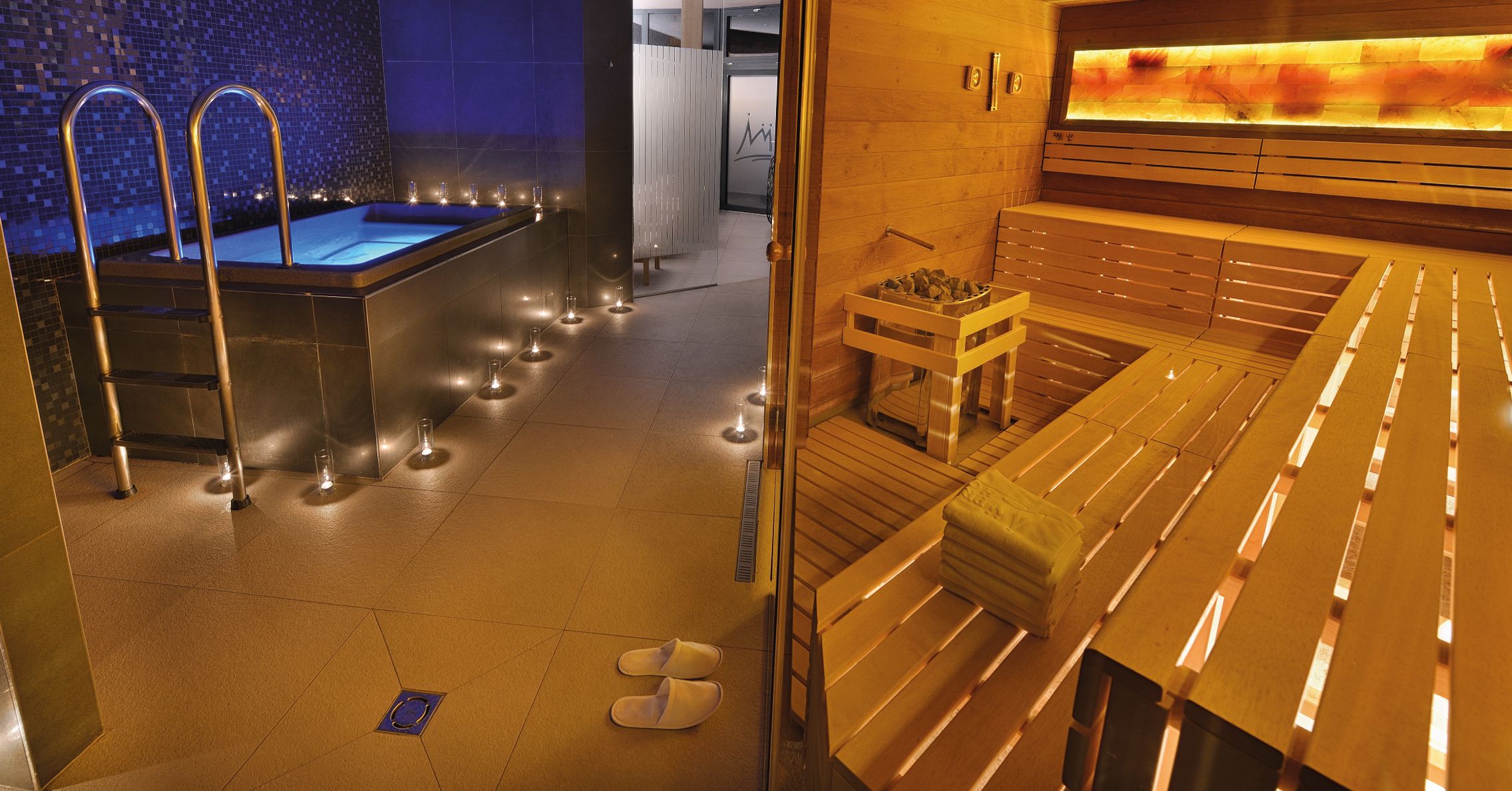 A sauna area with benches and a small table, a glass-walled hot tub, and a tiled floor with candles.