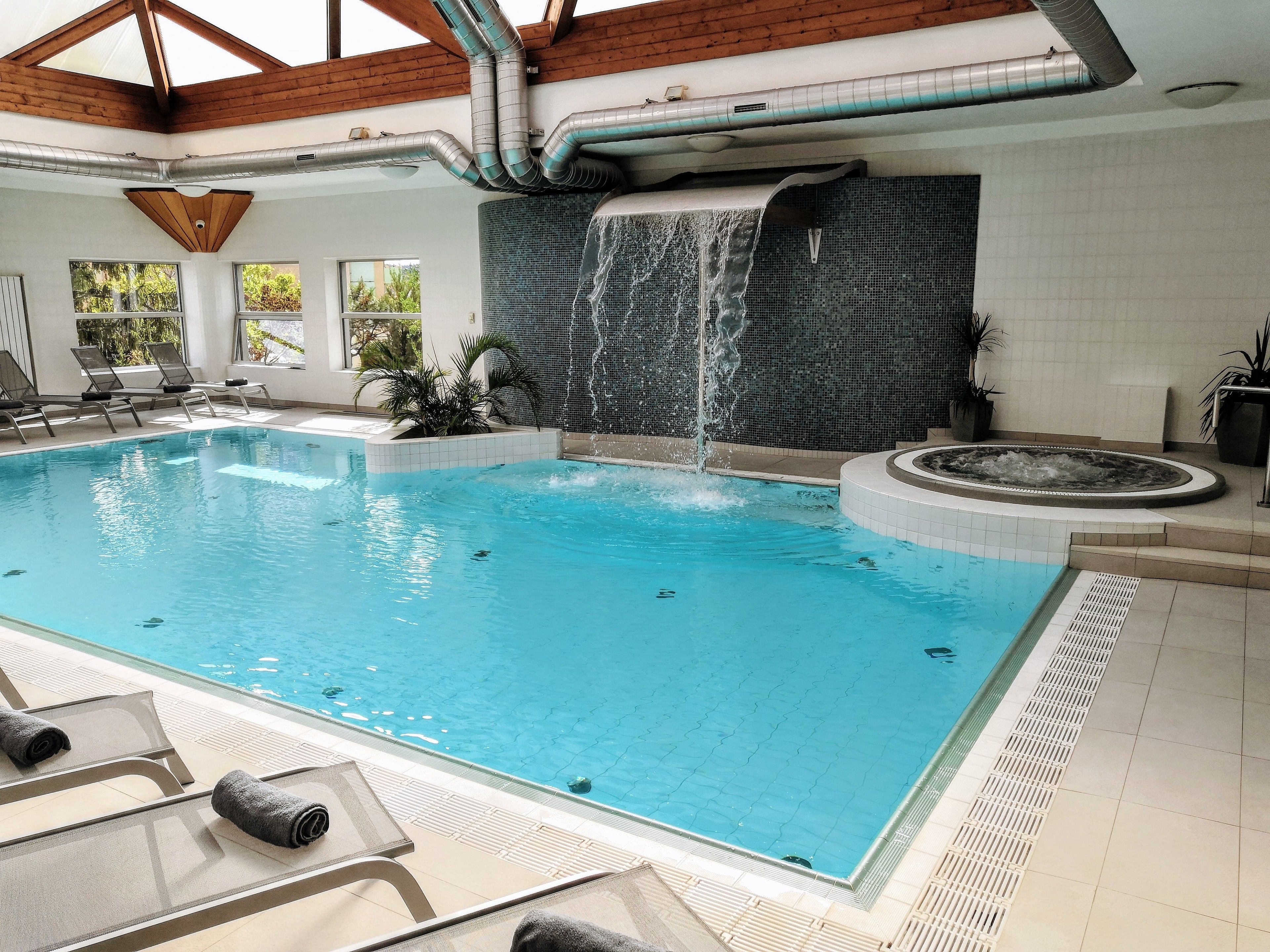 An indoor swimming pool with a waterfall feature, lounge chairs, and a tiled floor.