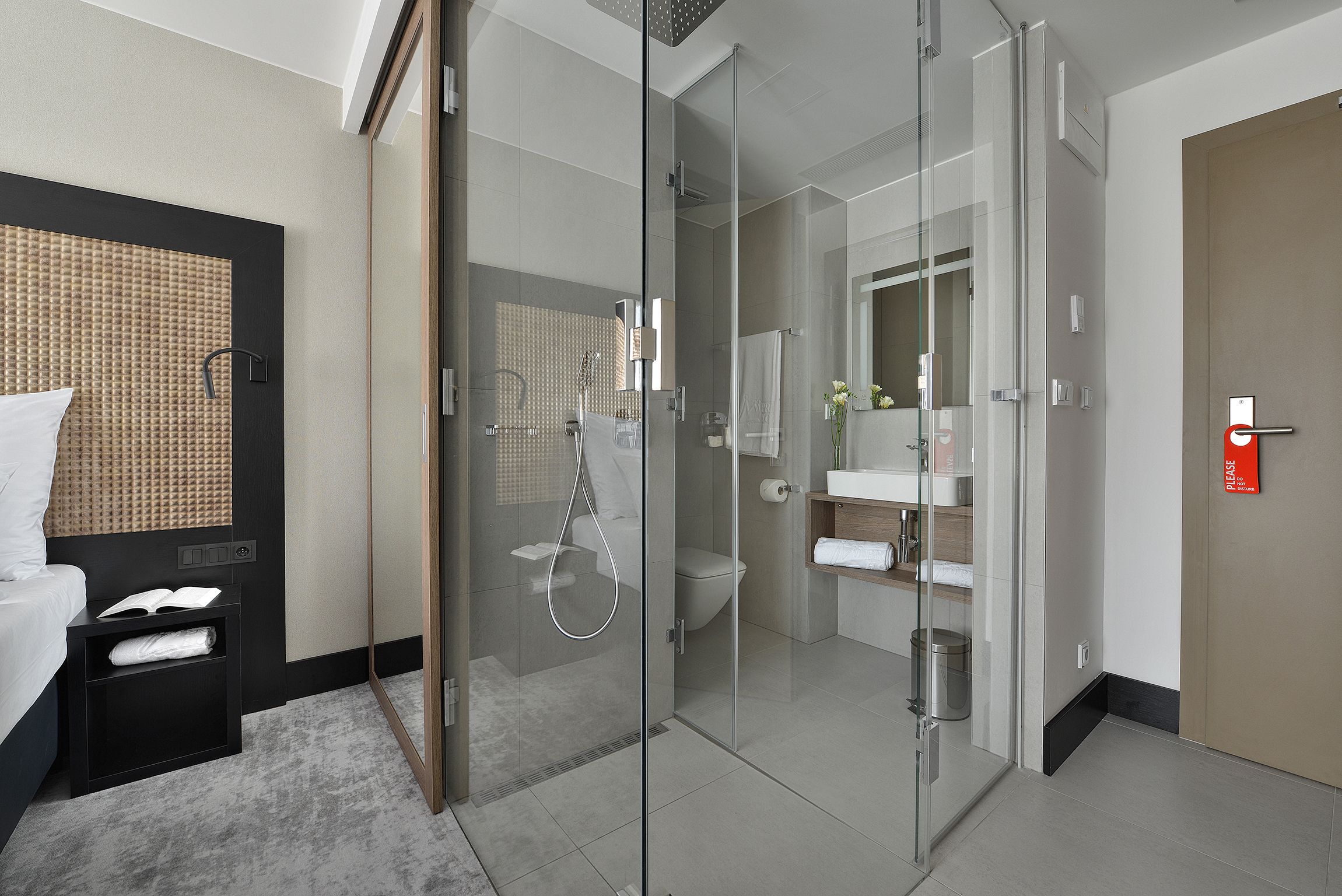 A modern hotel room with a glass-enclosed shower, a bed with a headboard, a bedside table with a book and a towel, and a bathroom with a toilet, sink, and mirror.