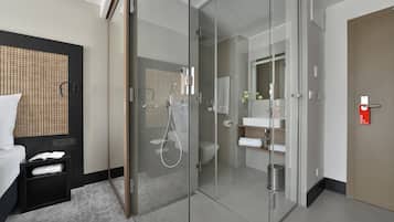 A modern hotel room with a glass-enclosed shower, a bed with a headboard, a bedside table with a book and a towel, and a bathroom with a toilet, sink, and mirror.