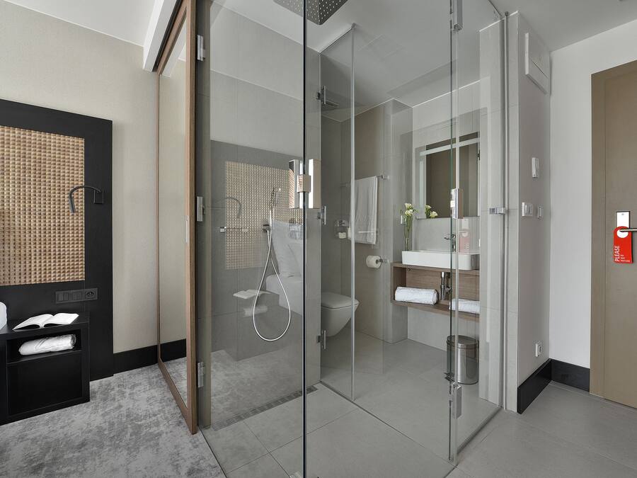 A modern hotel room with a glass-enclosed shower, a bed with a headboard, a bedside table with a book and a towel, and a bathroom with a toilet, sink, and mirror.