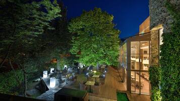An outdoor dining area with tables and chairs, surrounded by trees and a stone building.