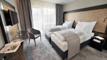 Habitación de hotel con dos camas, televisor montado en la pared, escritorio con silla y una mesita con un libro y gafas.
