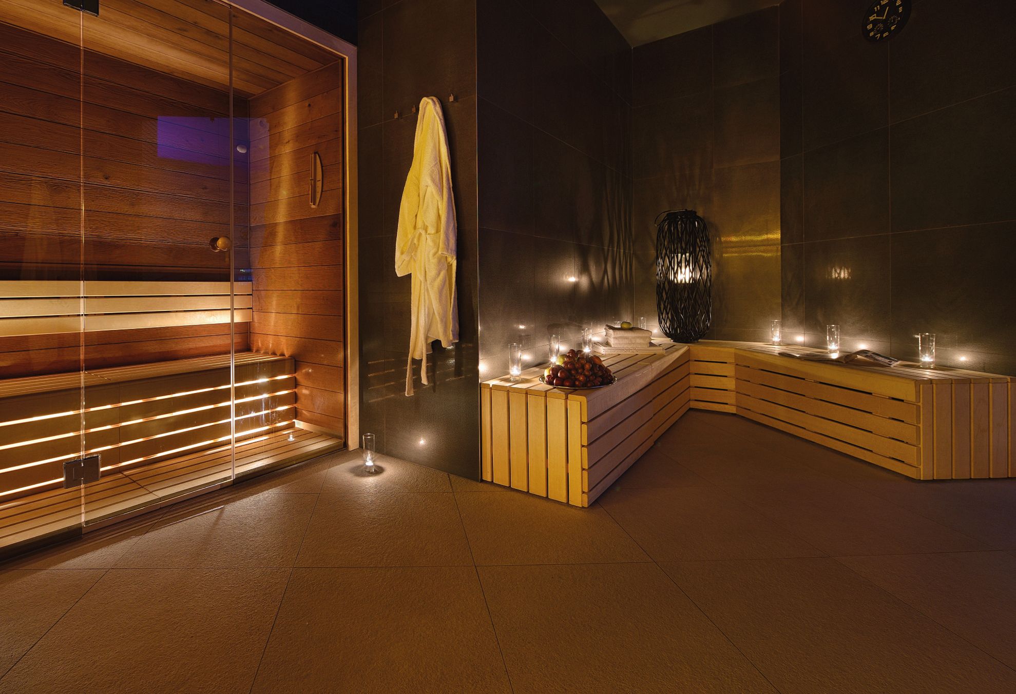 Un spa moderno con sauna de madera, sala de vapor con paredes de vidrio y zona de estar con velas y frutas.